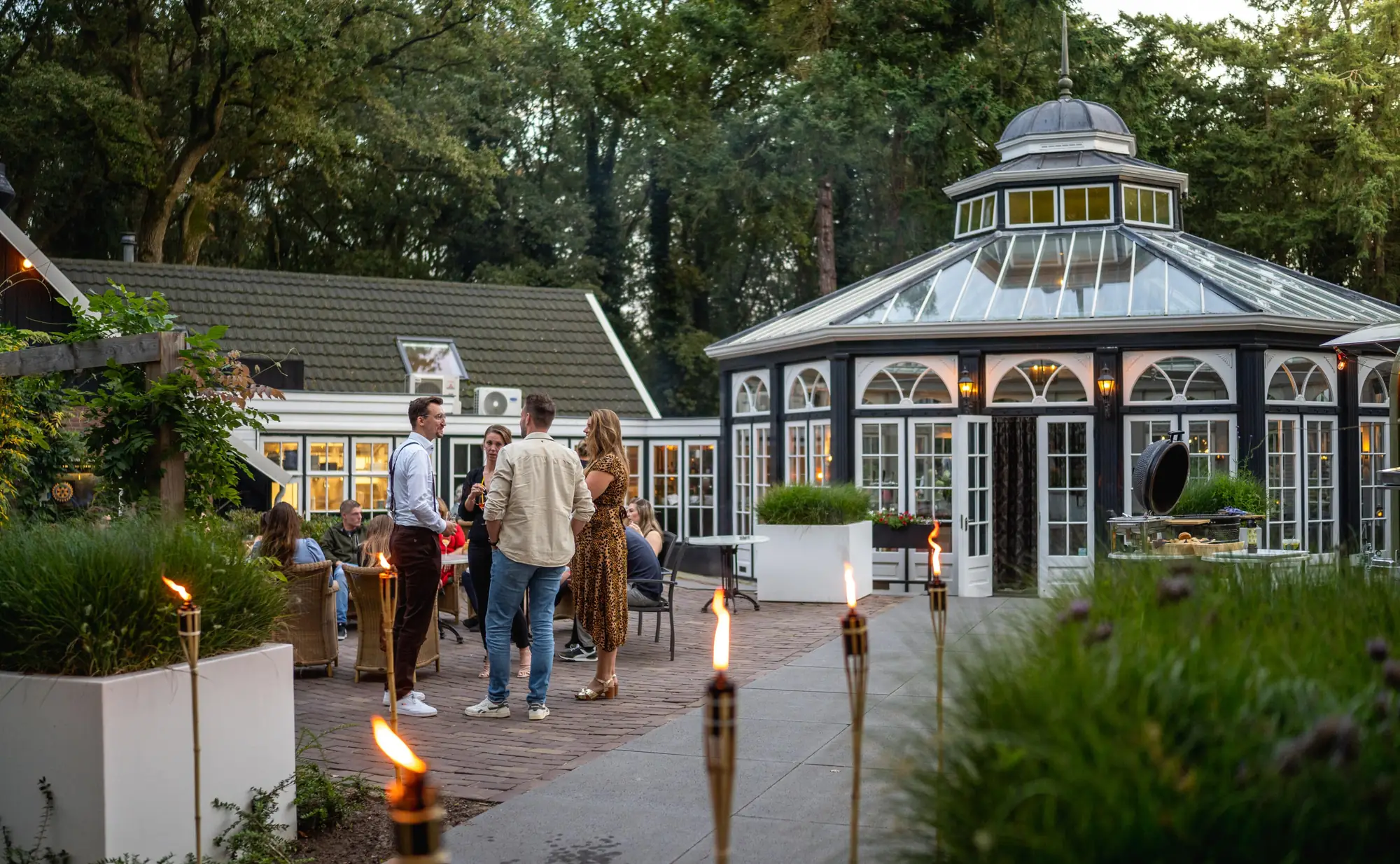 Gasten met een drankje op het terras van de Agnietenberg in de schemer, met fakkels op de voorgrond en de verlichte Wintertuin erachter