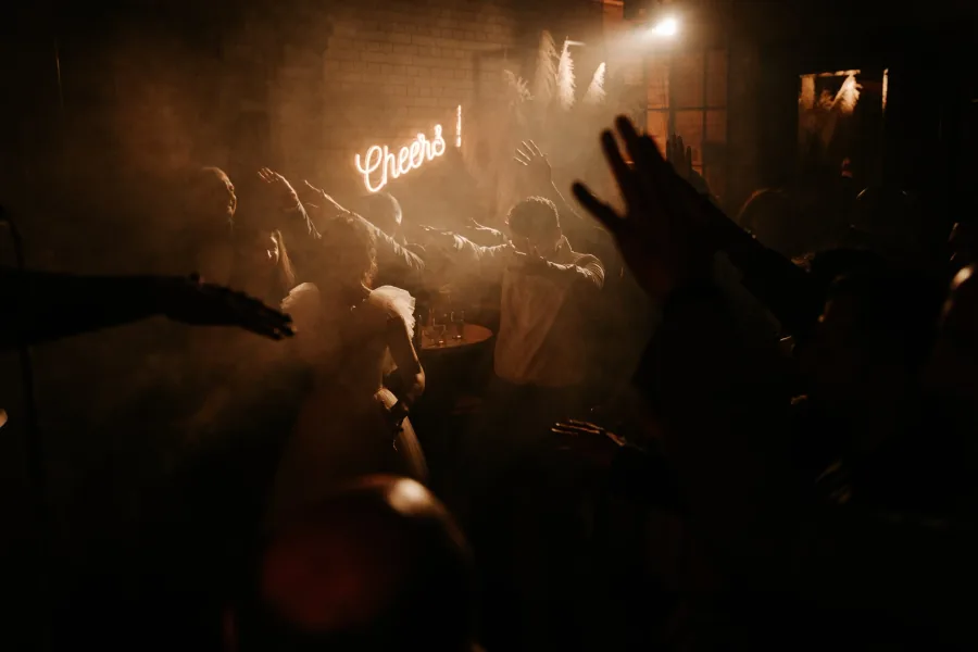 Handen in de lucht op de dansvloer, met een neonbord waarop Cheers staat