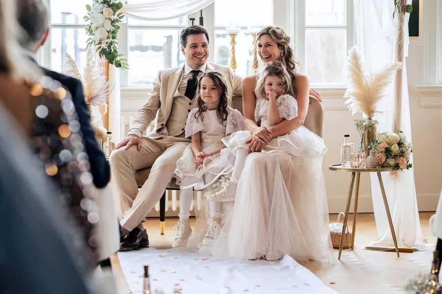 Een bruidspaar zit met twee kinderen in een lichte zaal met bloemen en droogbloemen